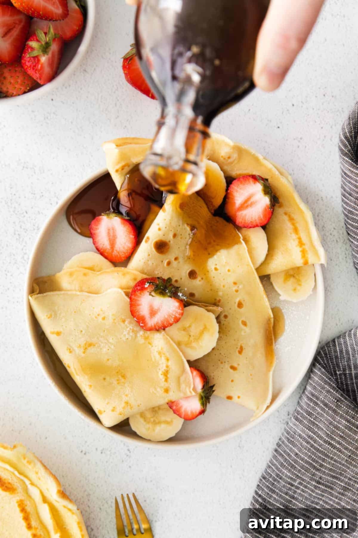 A stack of freshly made crepes being drizzled with rich, golden maple syrup, showcasing their thin, delicate texture.