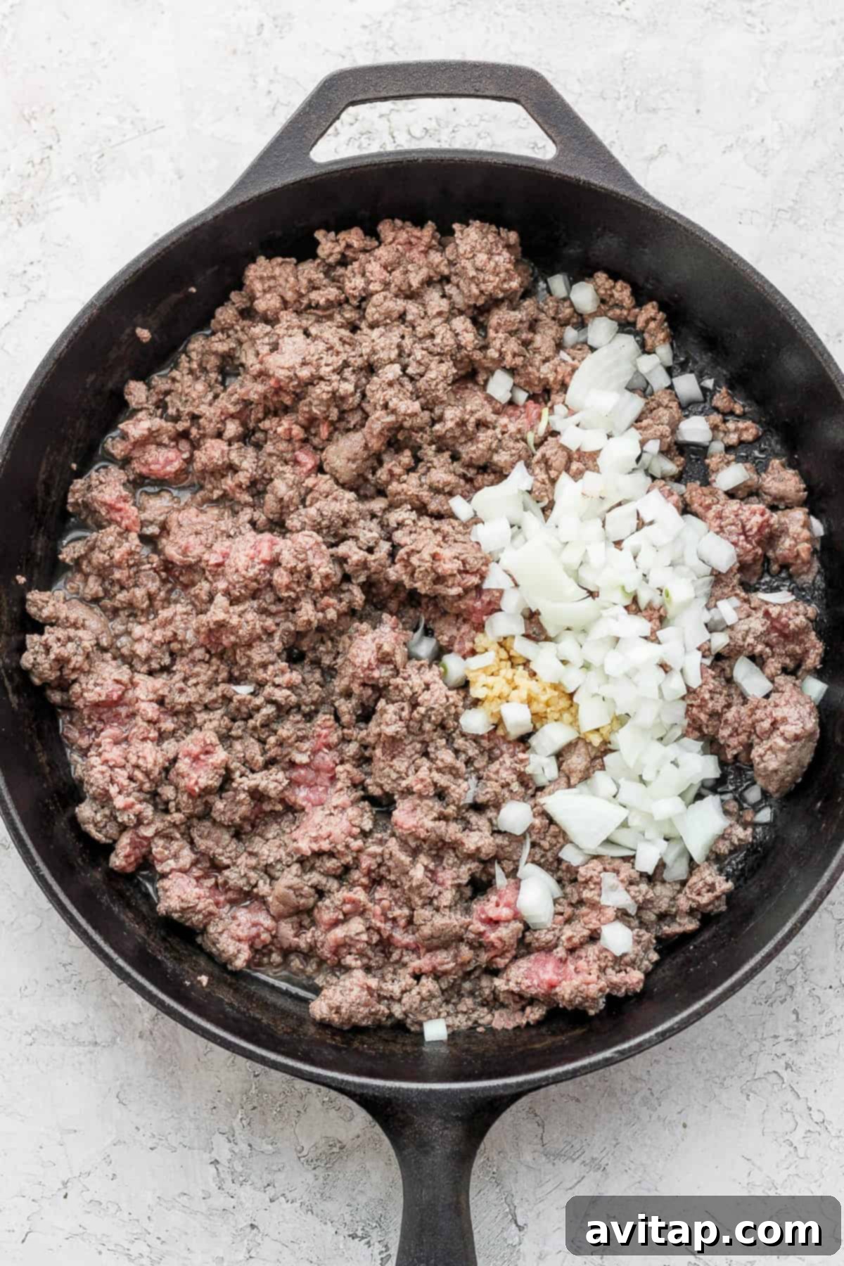 Ground beef cooking in a skillet with finely diced yellow onion and minced garlic, releasing aromatic flavors.