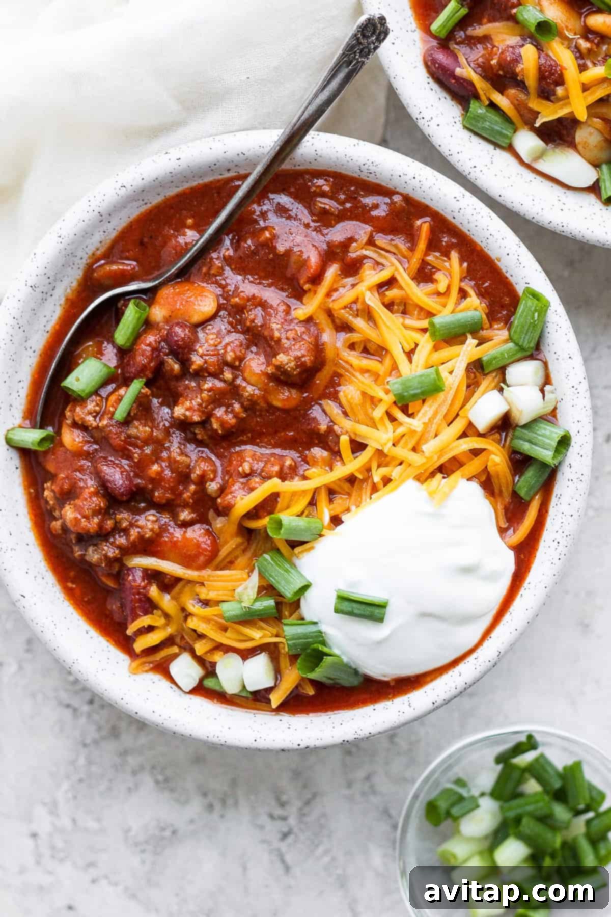 A hearty bowl of slow cooker chili topped generously with cool sour cream, shredded cheddar cheese, and vibrant diced green onions.