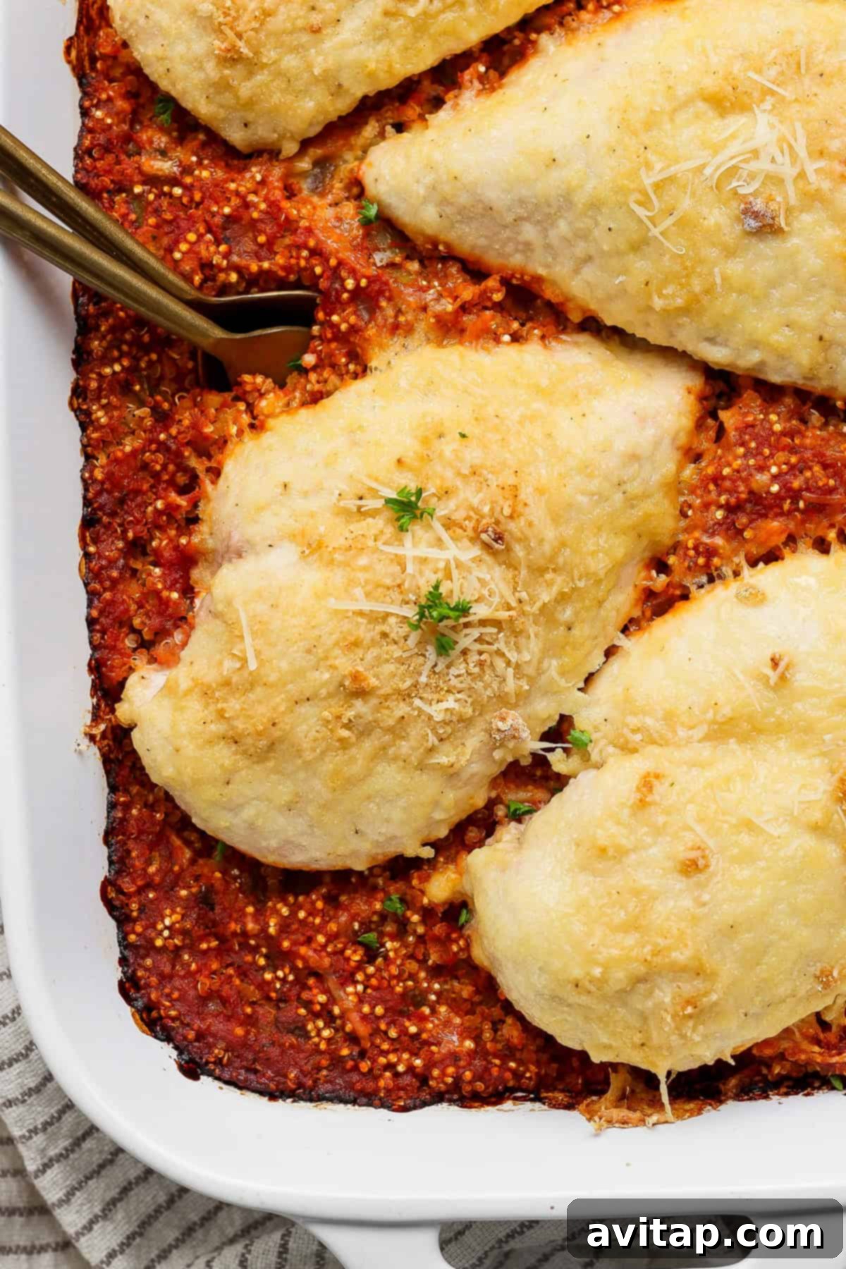Chicken Parmesan Quinoa Bake 2 A close-up shot of a baked chicken parmesan quinoa casserole in a white baking dish, topped with melted cheese and fresh herbs, ready to be served. The rich red quinoa base is visible beneath the golden-brown chicken.