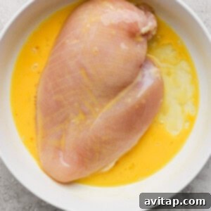Raw chicken breast in a white bowl with beaten eggs on a gray textured surface.