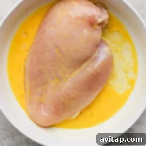 Raw chicken breast in a white bowl with beaten eggs on a gray textured surface.