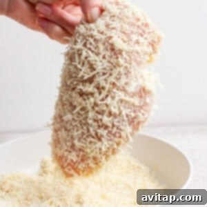 A hand holding a raw chicken breast coated with grated cheese over a white bowl.