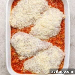 Four uncooked chicken breasts covered in grated cheese are placed on top of a layer of tomato sauce in a white baking dish.
