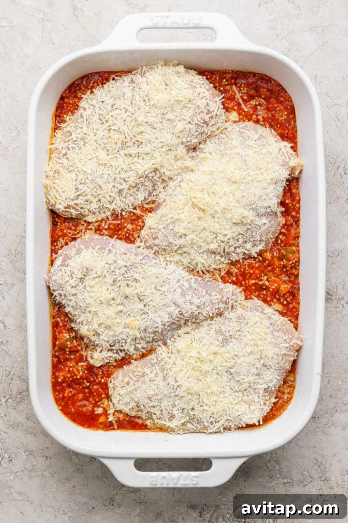 Chicken Parmesan Quinoa Bake 5 Four uncooked chicken breasts, generously coated in a parmesan and herb breading, are carefully arranged on top of a vibrant red marinara quinoa base in a white baking dish, ready for the oven.
