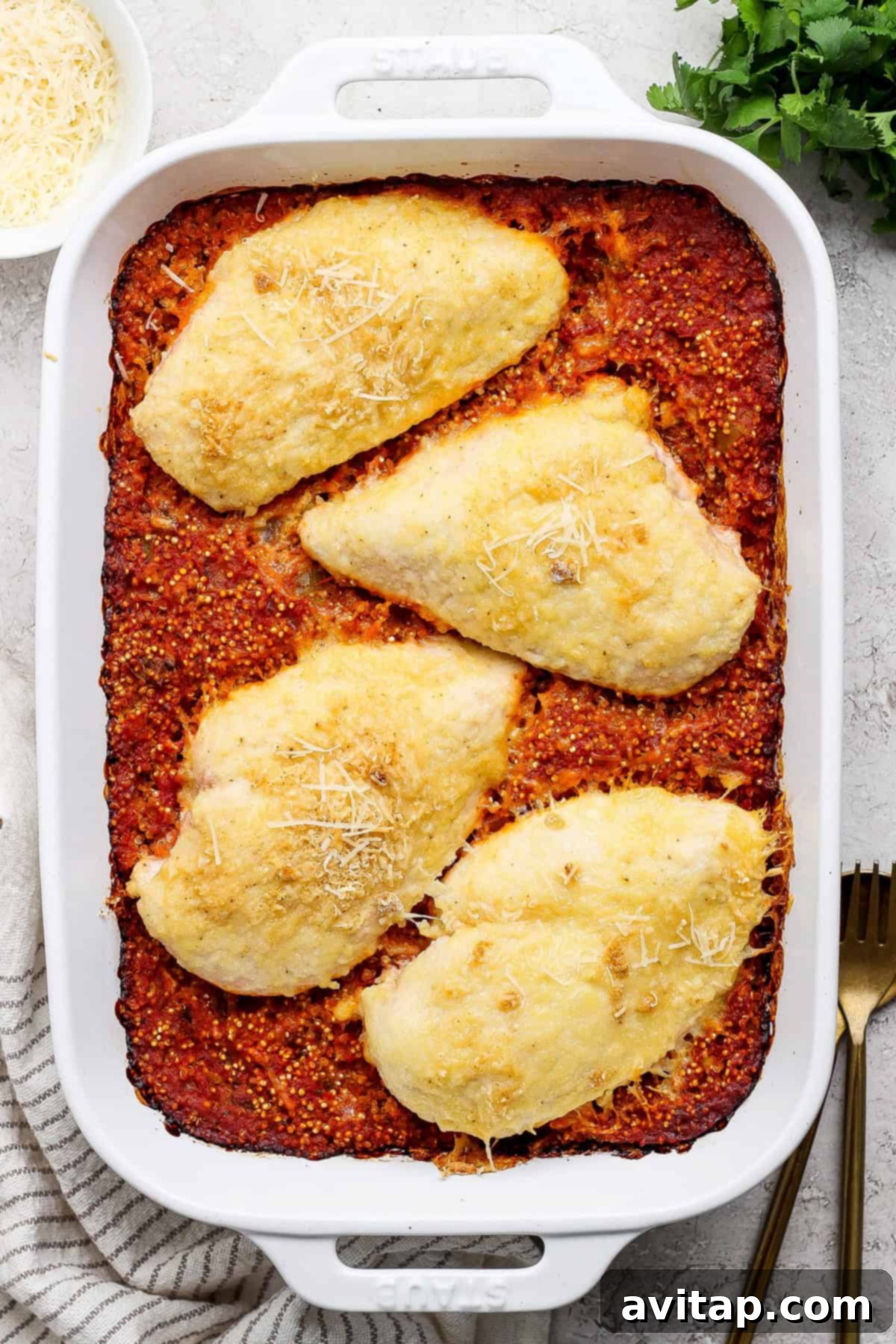 Parmesan Chicken Quinoa Bake 6 Beautifully baked chicken breasts topped with golden-brown cheese in a white casserole dish, resting on a vibrant red sauce base, with a fork and cloth napkin beside it, ready to be served.