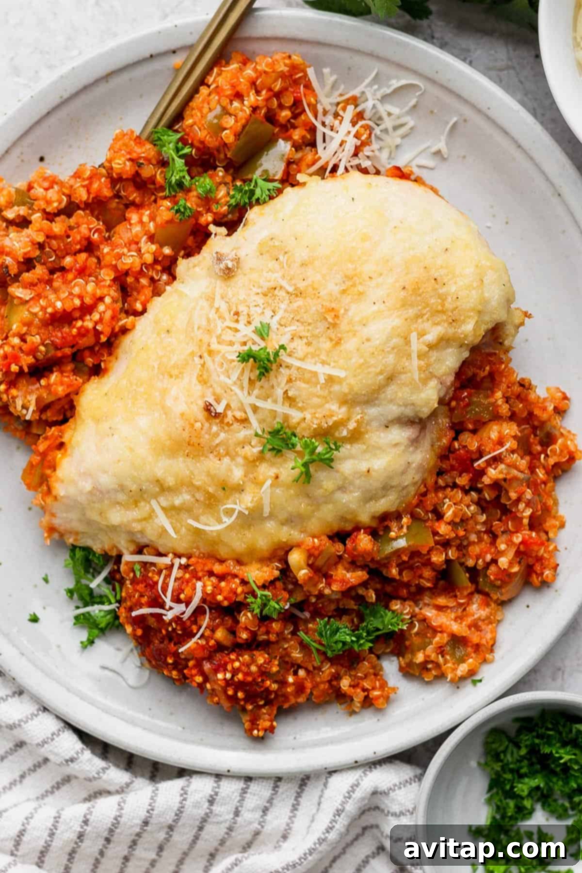 Chicken Parmesan Quinoa Bake 7 A single serving of chicken parmesan quinoa casserole, featuring a grilled chicken breast topped with cheese and fresh parsley, resting on a bed of red tomato quinoa on a white plate.