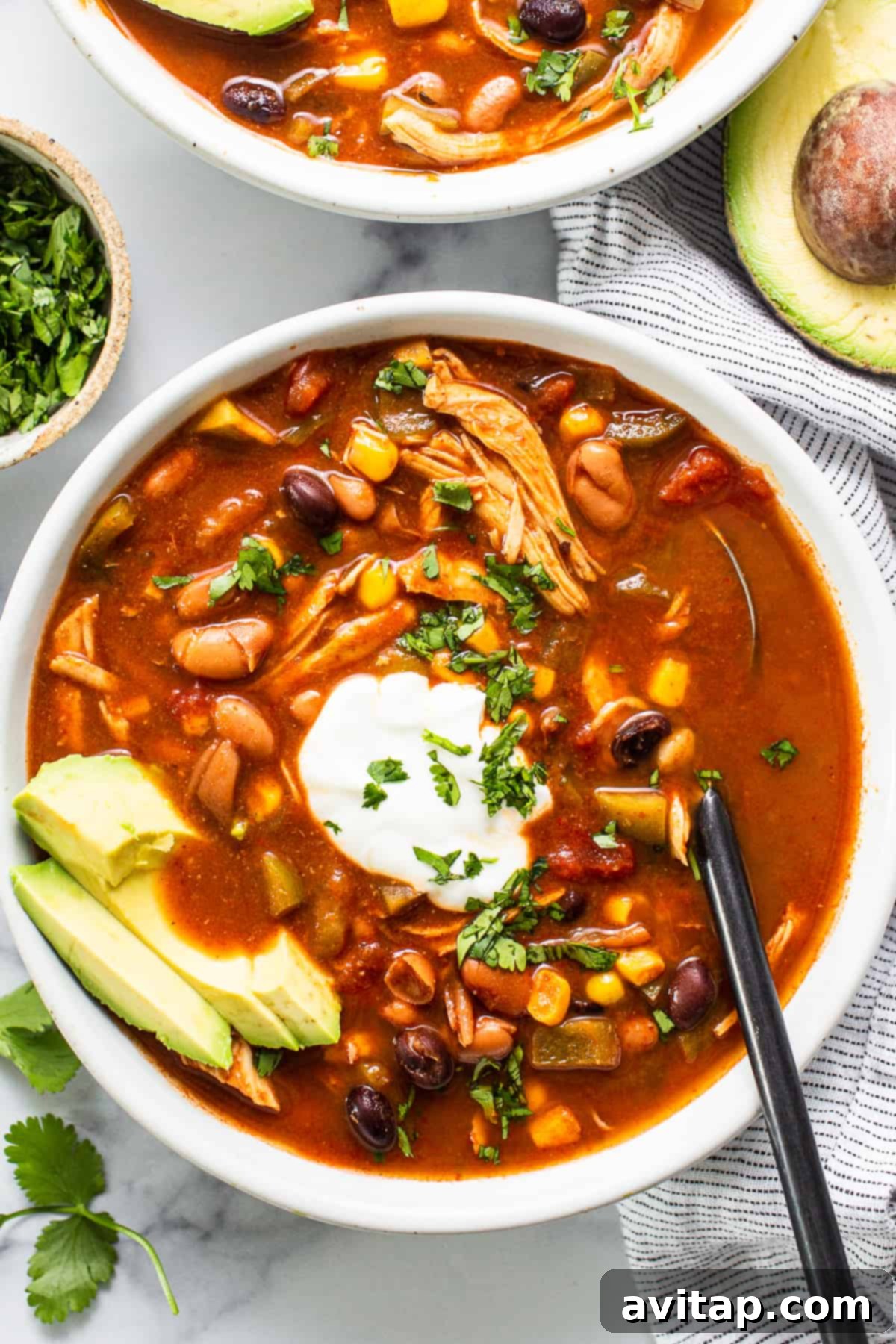 A large bowl of crockpot chicken enchilada soup topped with fresh avocado slices and a dollop of sour cream, garnished with cilantro, on a rustic wooden surface, with a slow cooker visible in the background, exuding warmth and comfort.