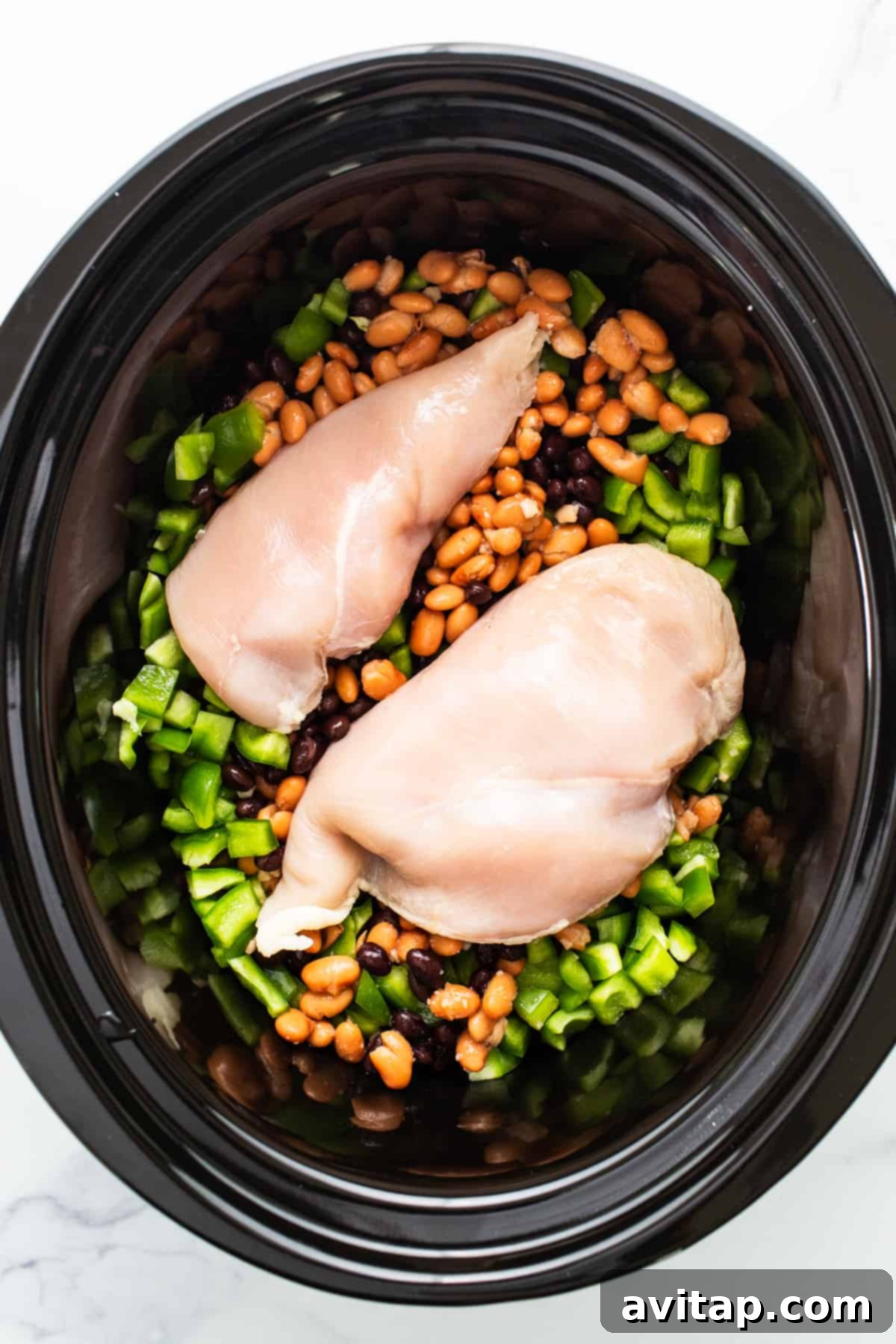 Close-up of a slow cooker filled with uncooked chicken enchilada soup ingredients: raw chicken breasts, various beans, diced bell peppers, and other vegetables submerged in a rich, red sauce, perfectly prepped and ready to begin cooking.