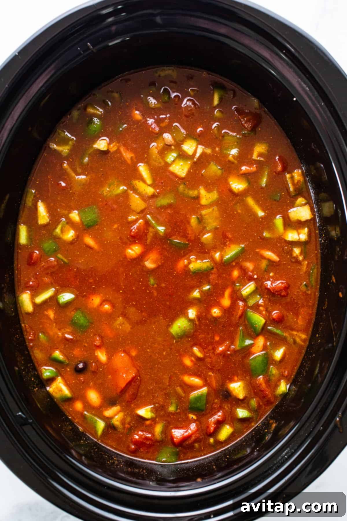 A close-up shot of the finished crockpot chicken enchilada soup simmering gently in a slow cooker, with a wooden spoon stirring the rich, red broth, showcasing visible pieces of shredded chicken, tender beans, and sweet corn, ready to be served.