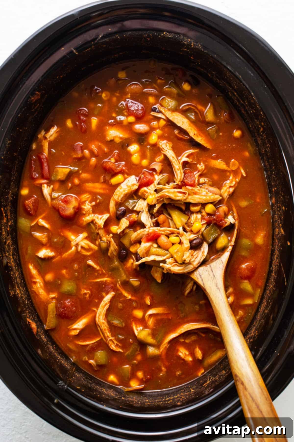 A close-up of a bowl of rich, red enchilada soup with shredded chicken, corn, and beans, beautifully plated and ready to be served, with a wooden ladle resting in the slow cooker in the background, signaling a delicious meal.
