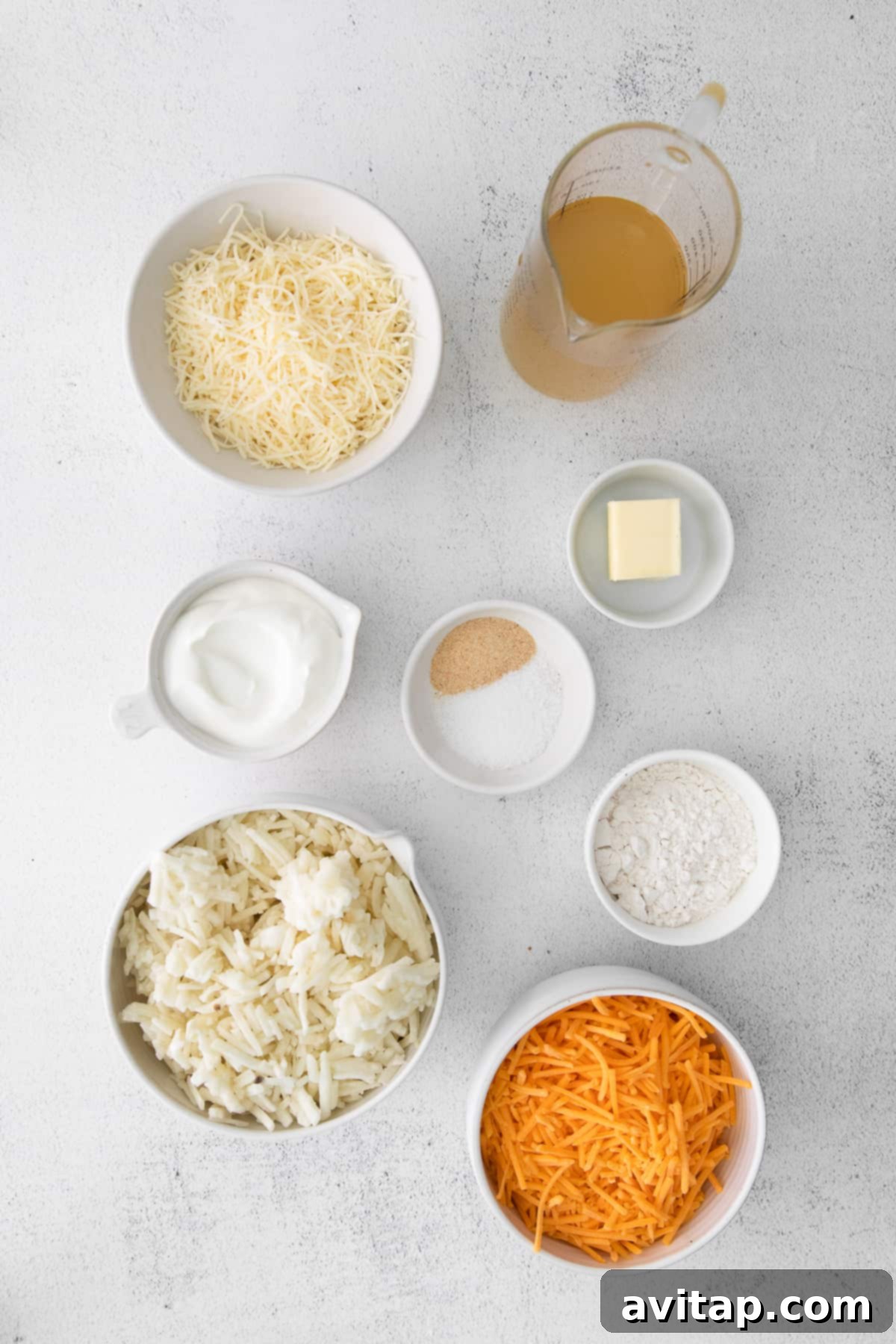 Fresh ingredients for homemade cheesy potato casserole laid out on a kitchen counter: hash browns, various cheeses, garlic powder, salt, butter, flour, broth, and sour cream.