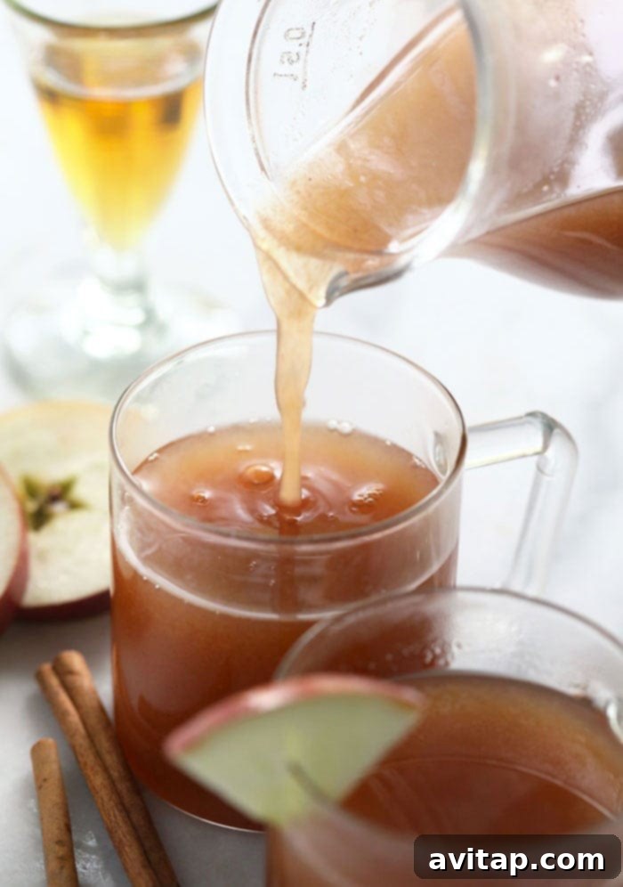 Warm spiked apple cider being poured into a mug, garnished with cinnamon sticks and orange slices.