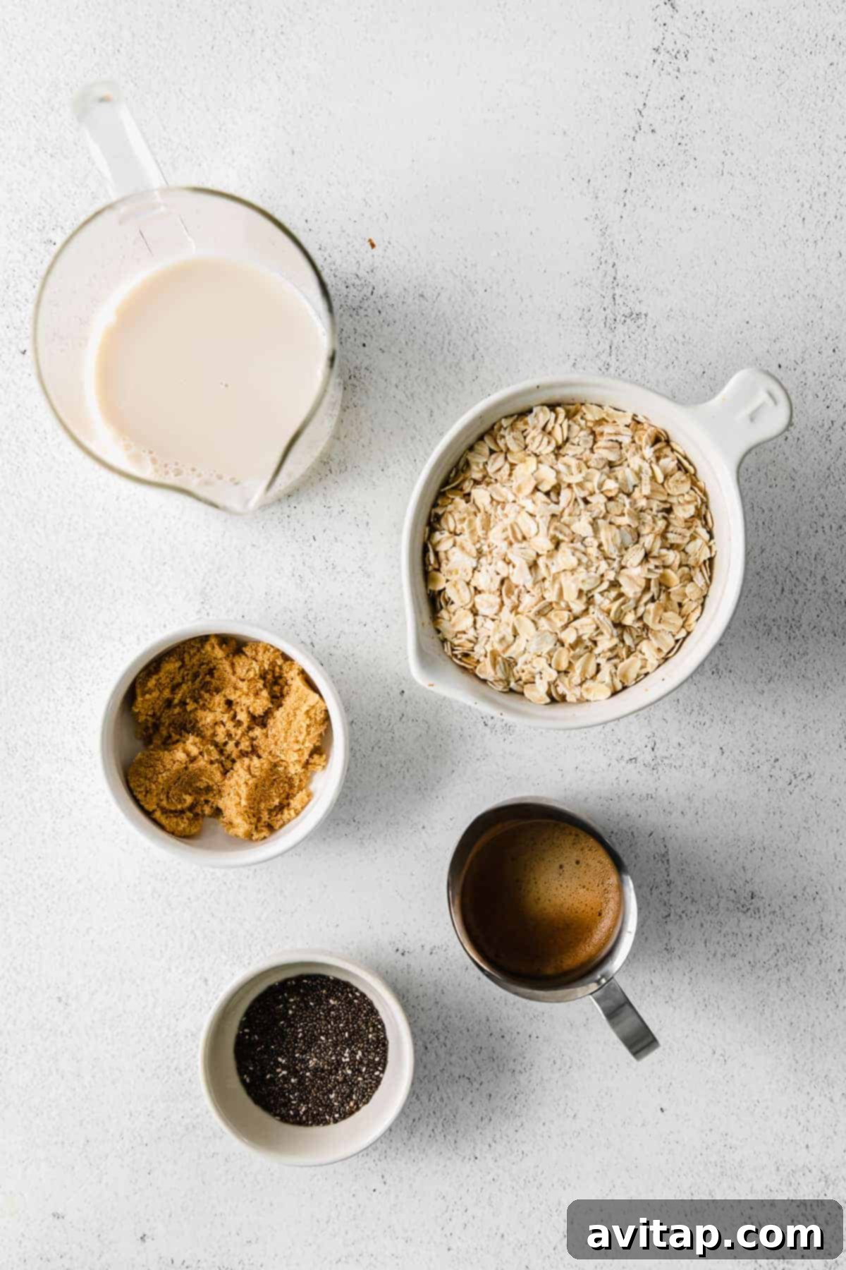 Assorted ingredients for overnight oats laid out neatly on a kitchen counter: rolled oats, dark brown sugar, chia seeds, a bottle of almond milk, and a container of espresso concentrate.