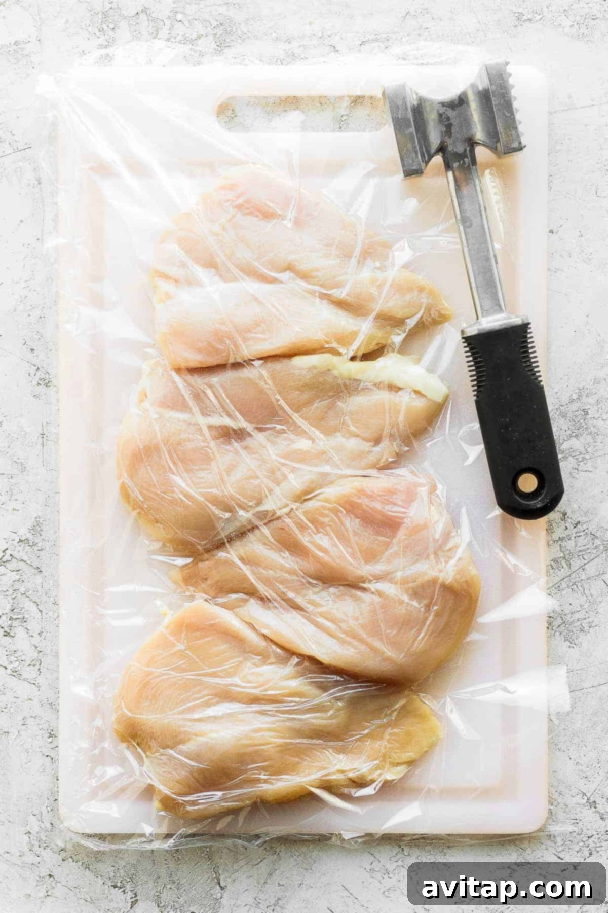 Raw chicken breasts on a cutting board, ready for tenderizing with a meat mallet.