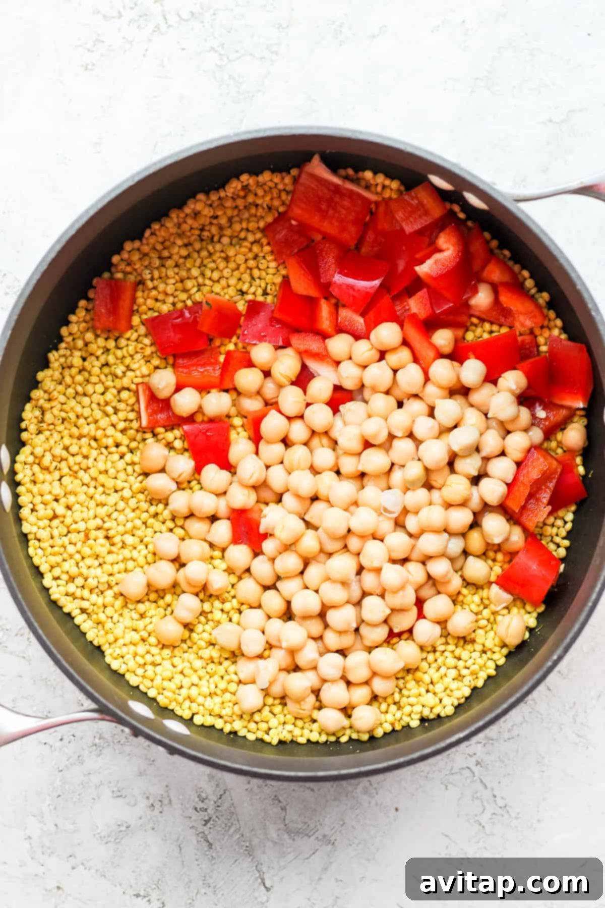 Red bell peppers, chickpeas, and lemon juice being added to toasted Israeli couscous in a pot, ready for simmering.