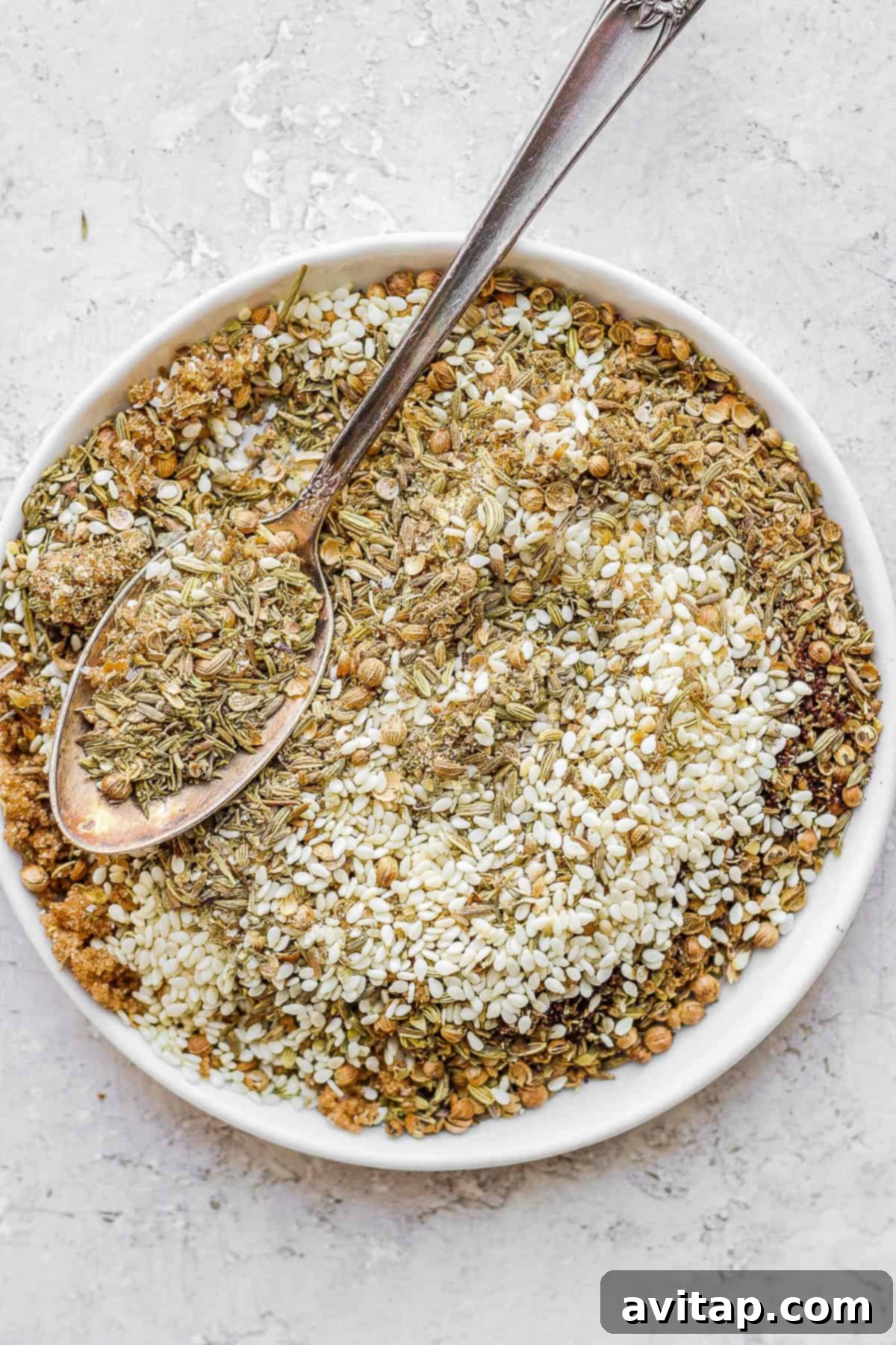 A close-up of freshly prepared za'atar seasoning on a rustic plate, next to a small wooden spoon, highlighting its vibrant green and red hues with white sesame seeds.