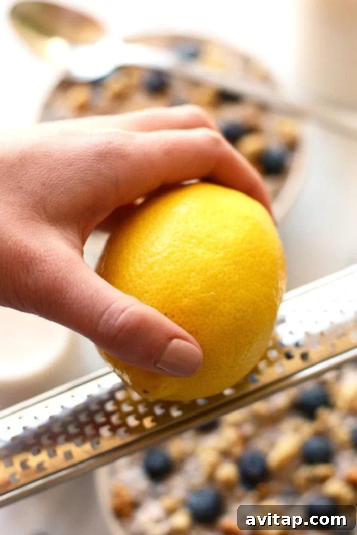 A vital ingredient: adding fresh lemon zest brightens the blueberry flavor in your overnight oats. Hands zesting a whole lemon over a bowl, illustrating the addition of fresh lemon zest to a recipe.