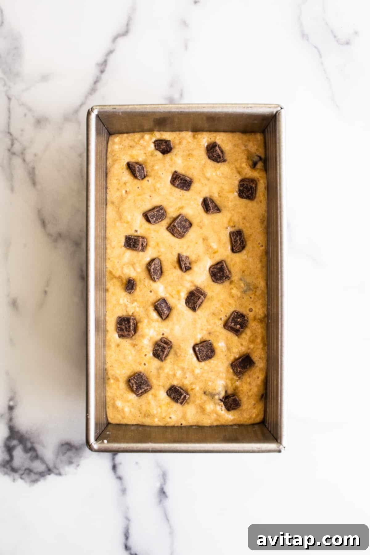 Thick banana bread batter swirled with dark chocolate chips, perfectly poured into a parchment-lined bread pan, ready for its journey into the oven.