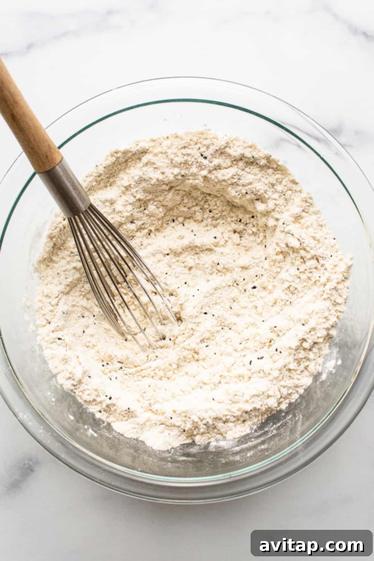 A glass bowl filled with dry ingredients for lemon poppy seed bread, including flour, baking powder, baking soda, poppy seeds, and lemon zest, ready to be mixed.