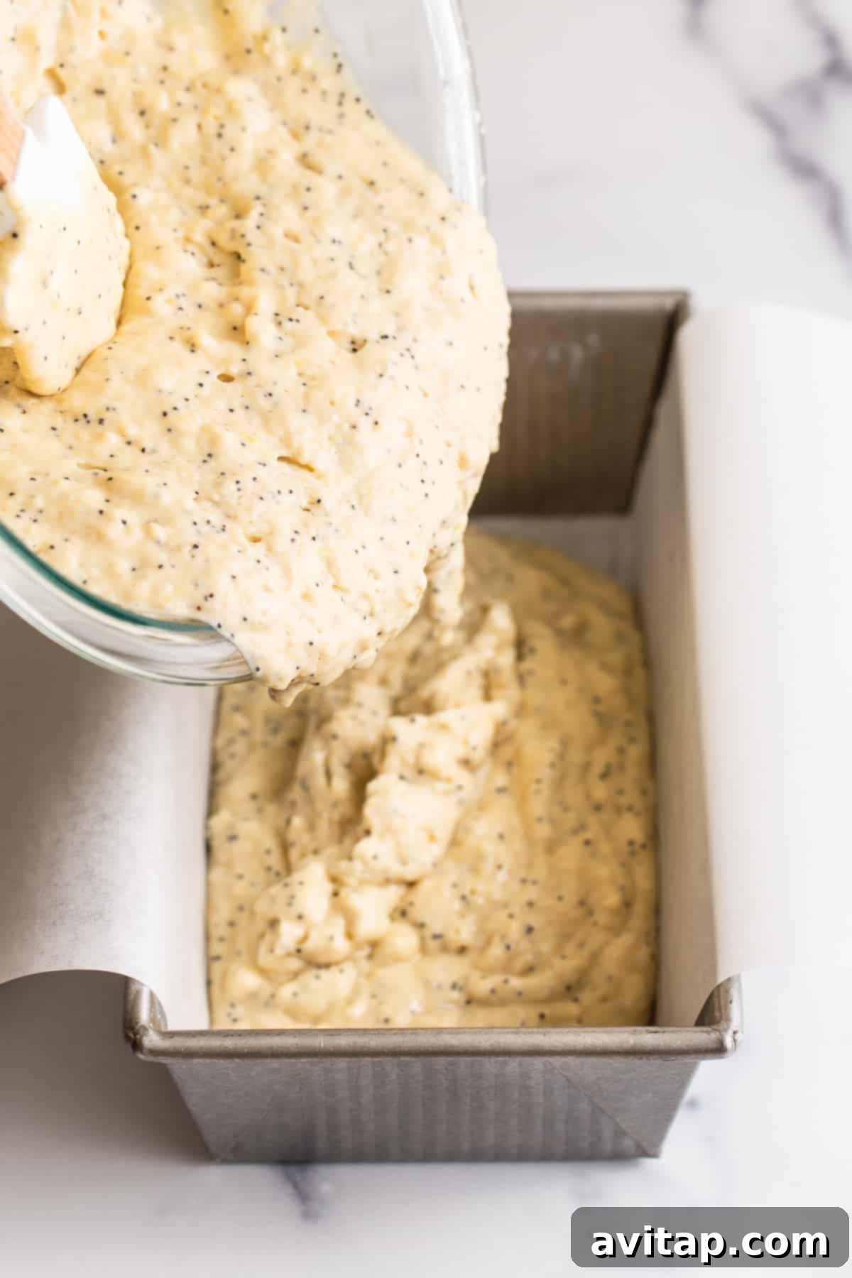 The lemon poppy seed bread batter being poured into a parchment-lined loaf pan, ready for baking.