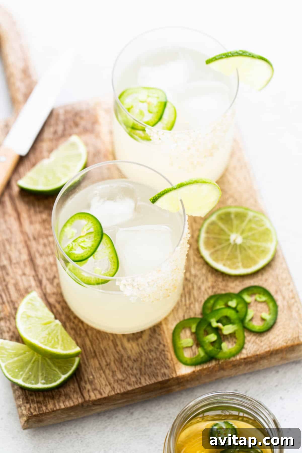 Two spicy mezcal margaritas, with lime and jalapeño garnishes, placed on a rustic cutting board.