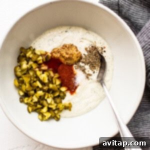 A bowl containing the creamy yogurt and cream cheese mixture, with minced pickles and spices added, ready for mixing.