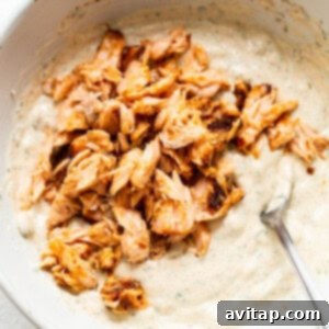 Flaked smoked salmon is gently folded into the seasoned creamy dip base in a white bowl, using a spoon.