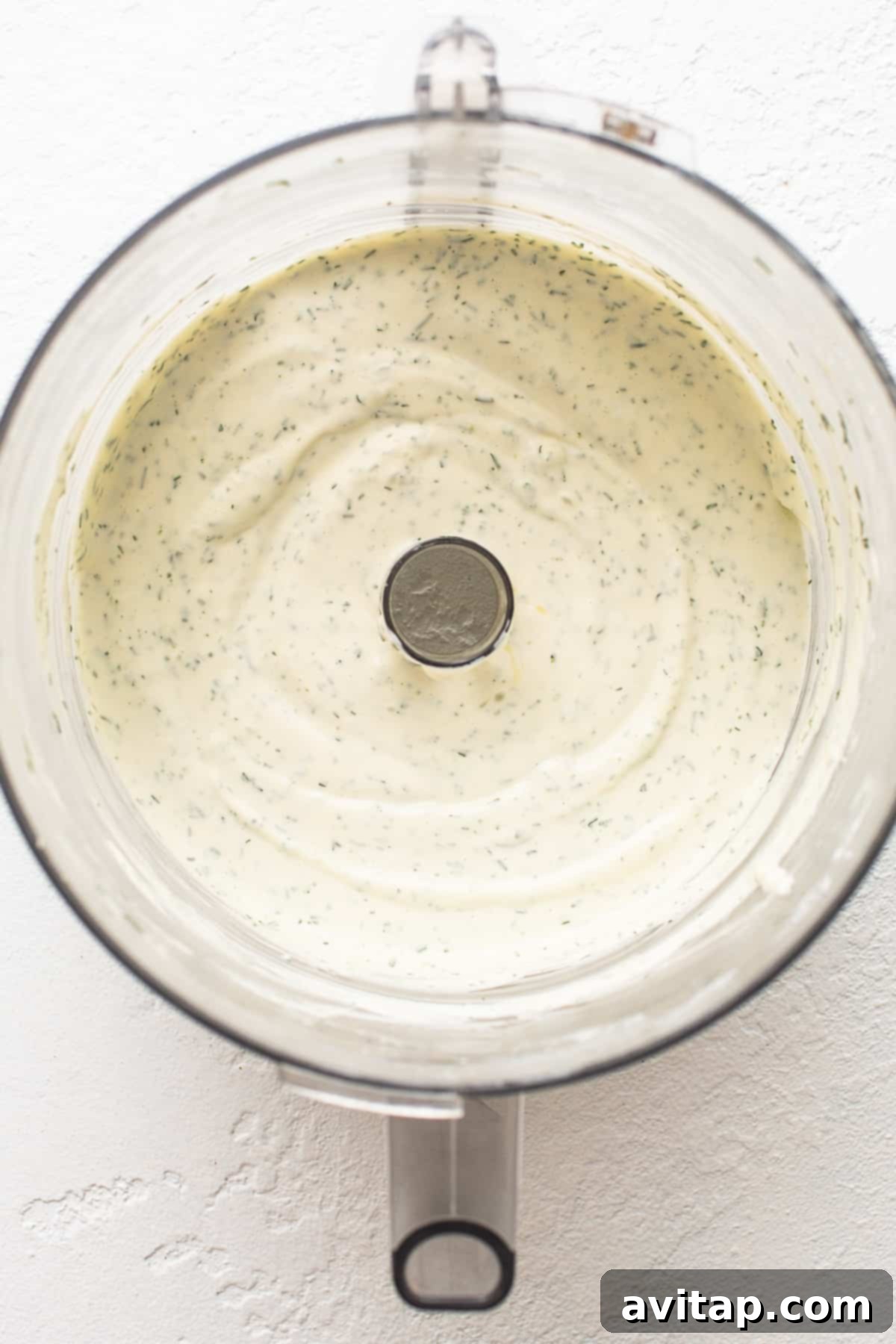A food processor bowl filled with a creamy, pale yellow mixture of Greek yogurt, cream cheese, olive oil, and lemon juice.