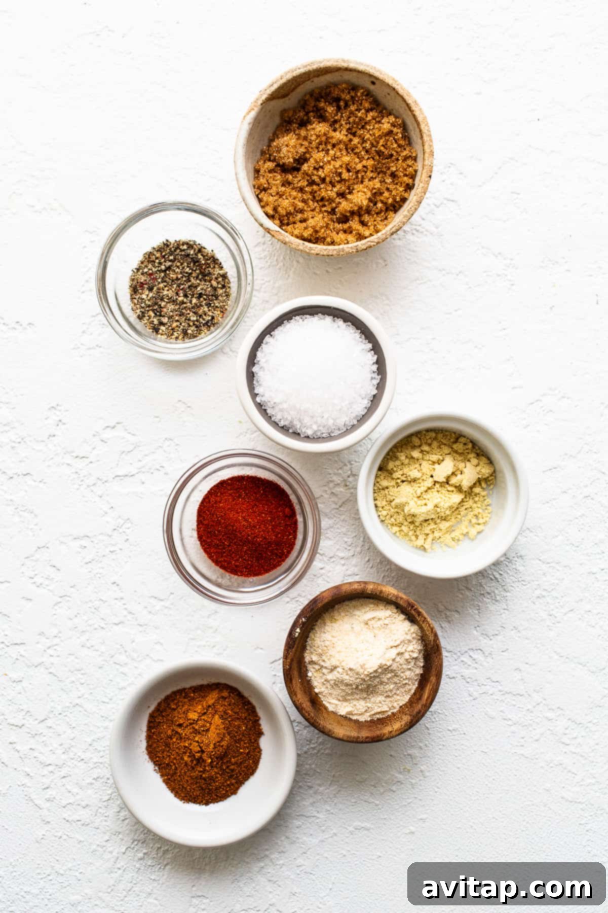 Tender Smoked Pork Shoulder 3 A vibrant display of dry rub ingredients, including dark brown sugar, coarse salt, ground black pepper, paprika, garlic powder, chili powder, and mustard powder, neatly arranged on a wooden surface.