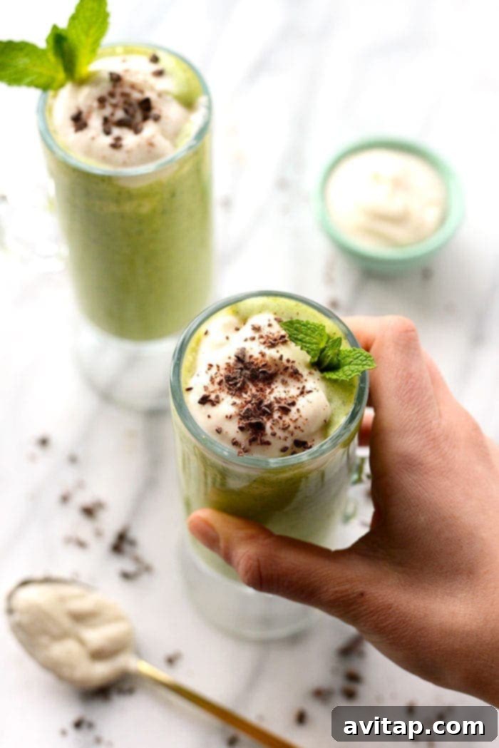 Hand holding a vibrant green Healthy Shamrock Shake, garnished with a sprinkle of dark chocolate chips, against a softly blurred background.