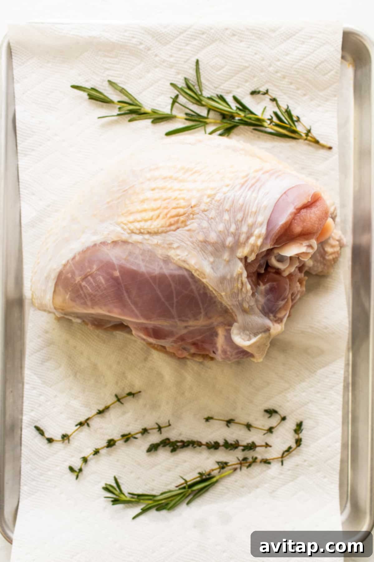 A brined turkey breast resting on a paper towel, ready for seasoning, with fresh herbs like rosemary and thyme visible nearby. 