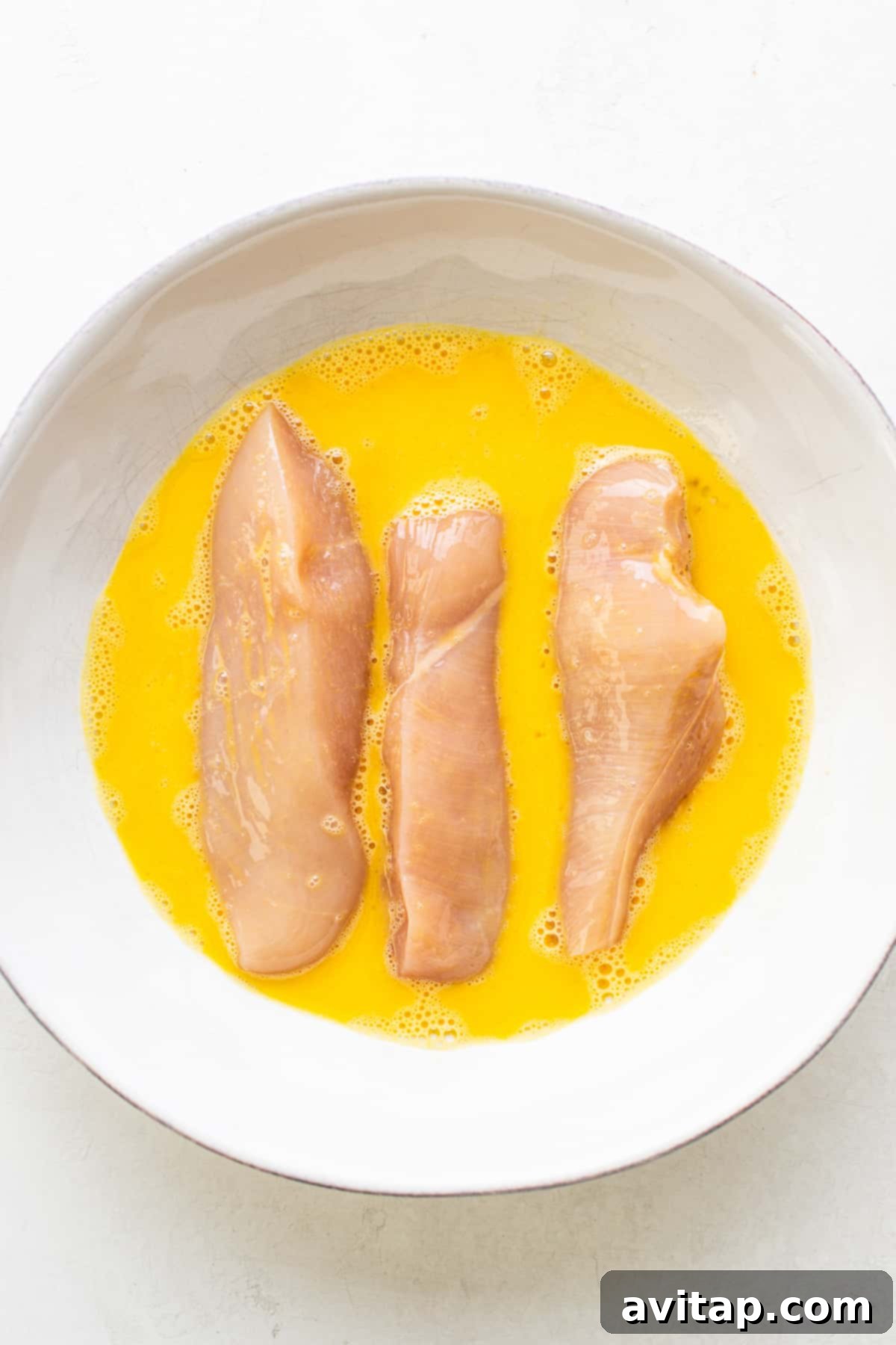 Raw chicken tenders being dipped in an egg wash, preparing them for breading.