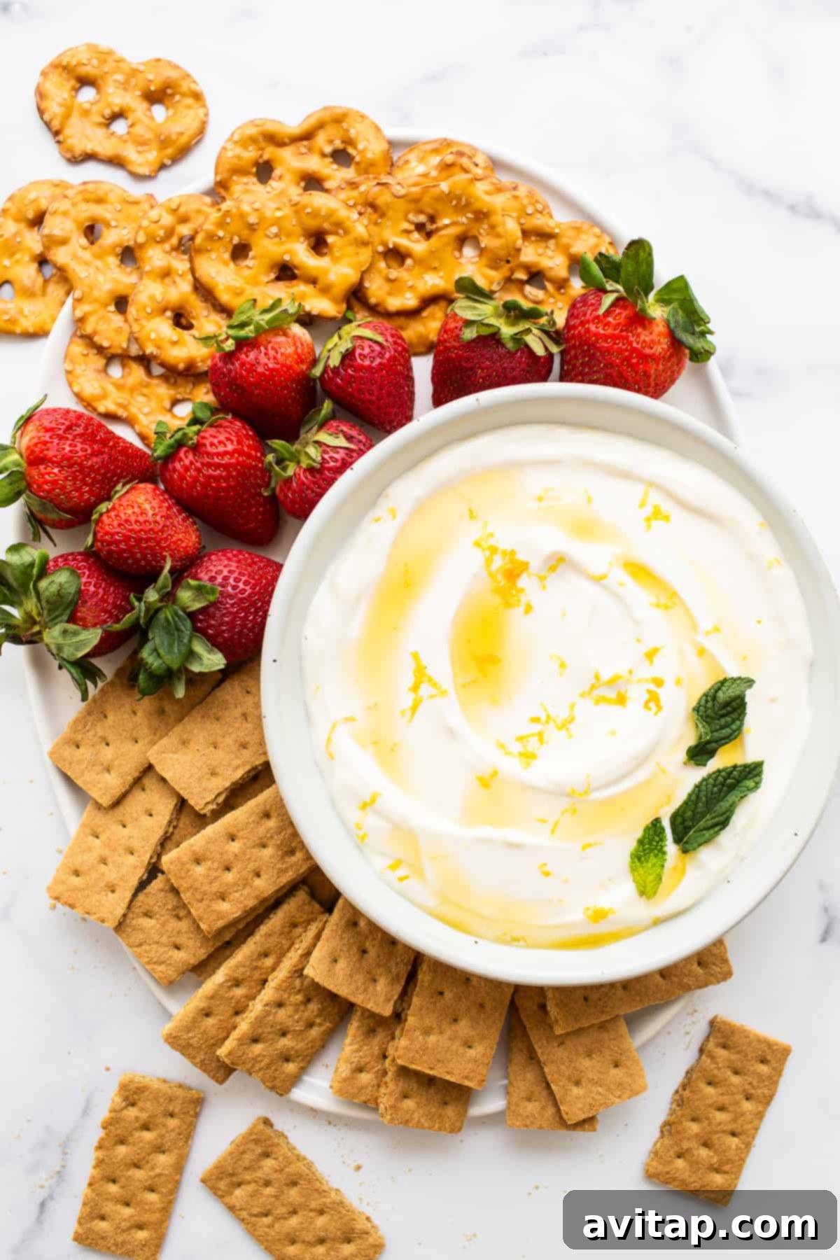 A vibrant platter featuring a central bowl of creamy Greek yogurt fruit dip, surrounded by an abundant assortment of fresh berries, crunchy pretzels, and golden graham crackers, perfect for sharing at any gathering.