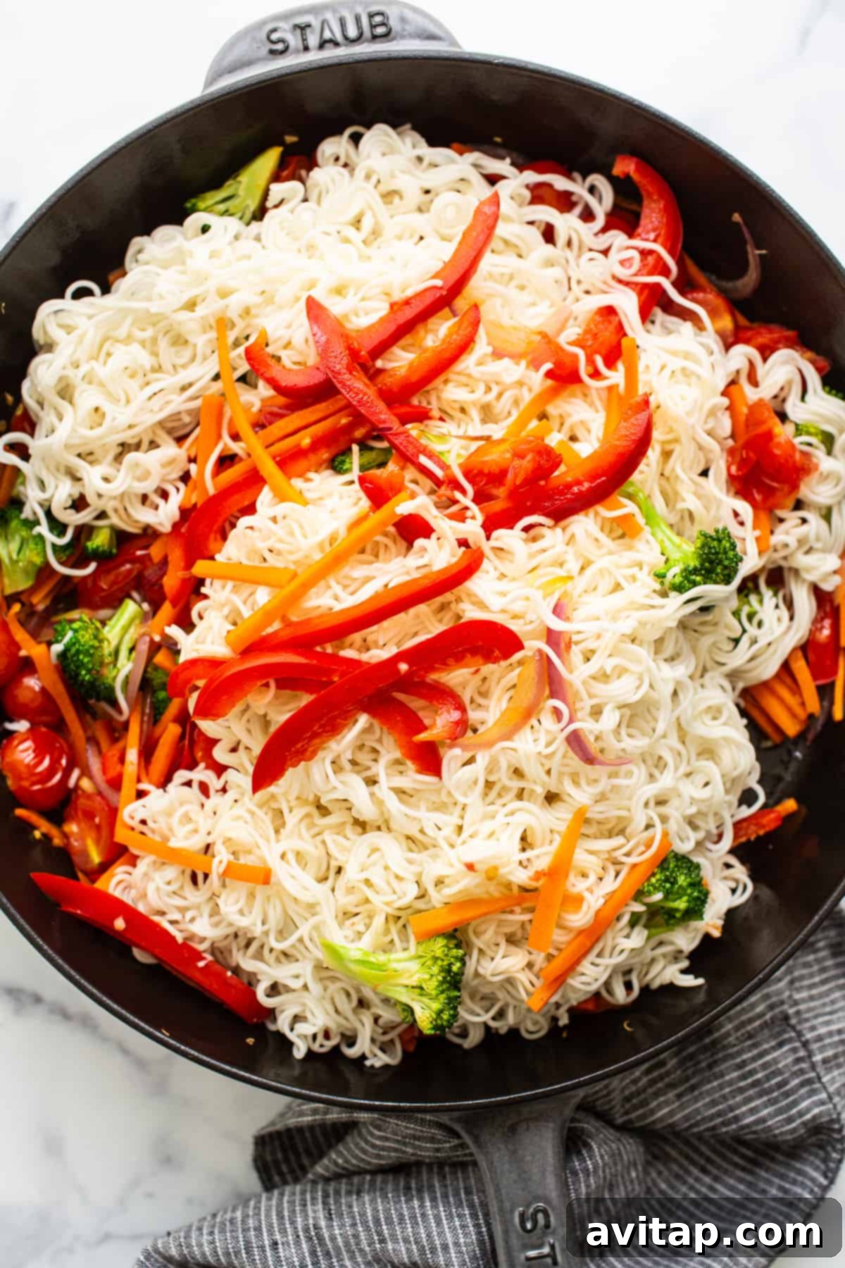 Cooked ramen noodles and sautéed vegetables being tossed together in a large skillet.
