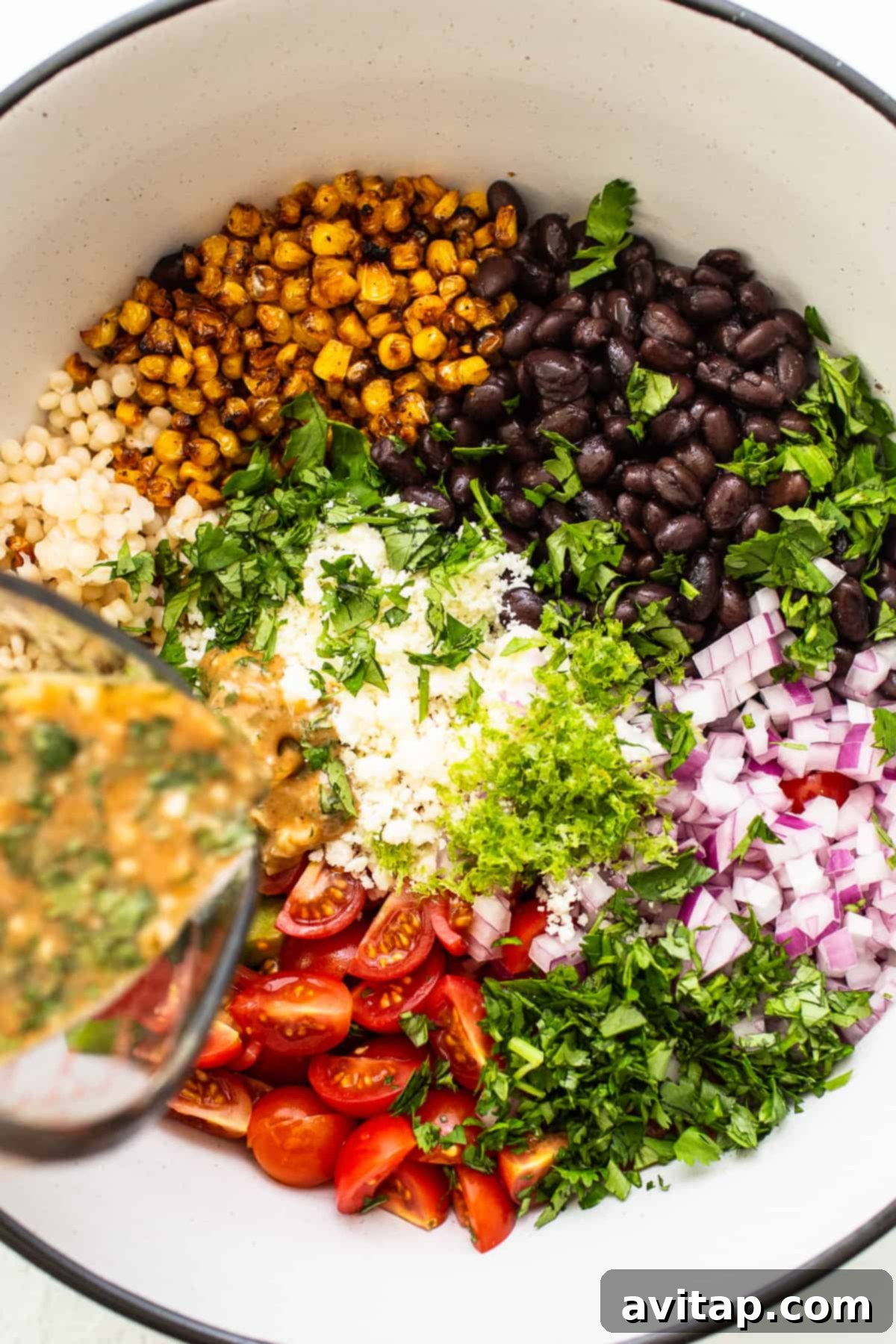 The final touch: pouring the vibrant cilantro lime dressing over the Black Bean Couscous Salad, ensuring every ingredient is perfectly coated. Close-up of cilantro lime dressing being poured over a colorful black bean couscous salad in a large bowl, highlighting the vibrant ingredients.
