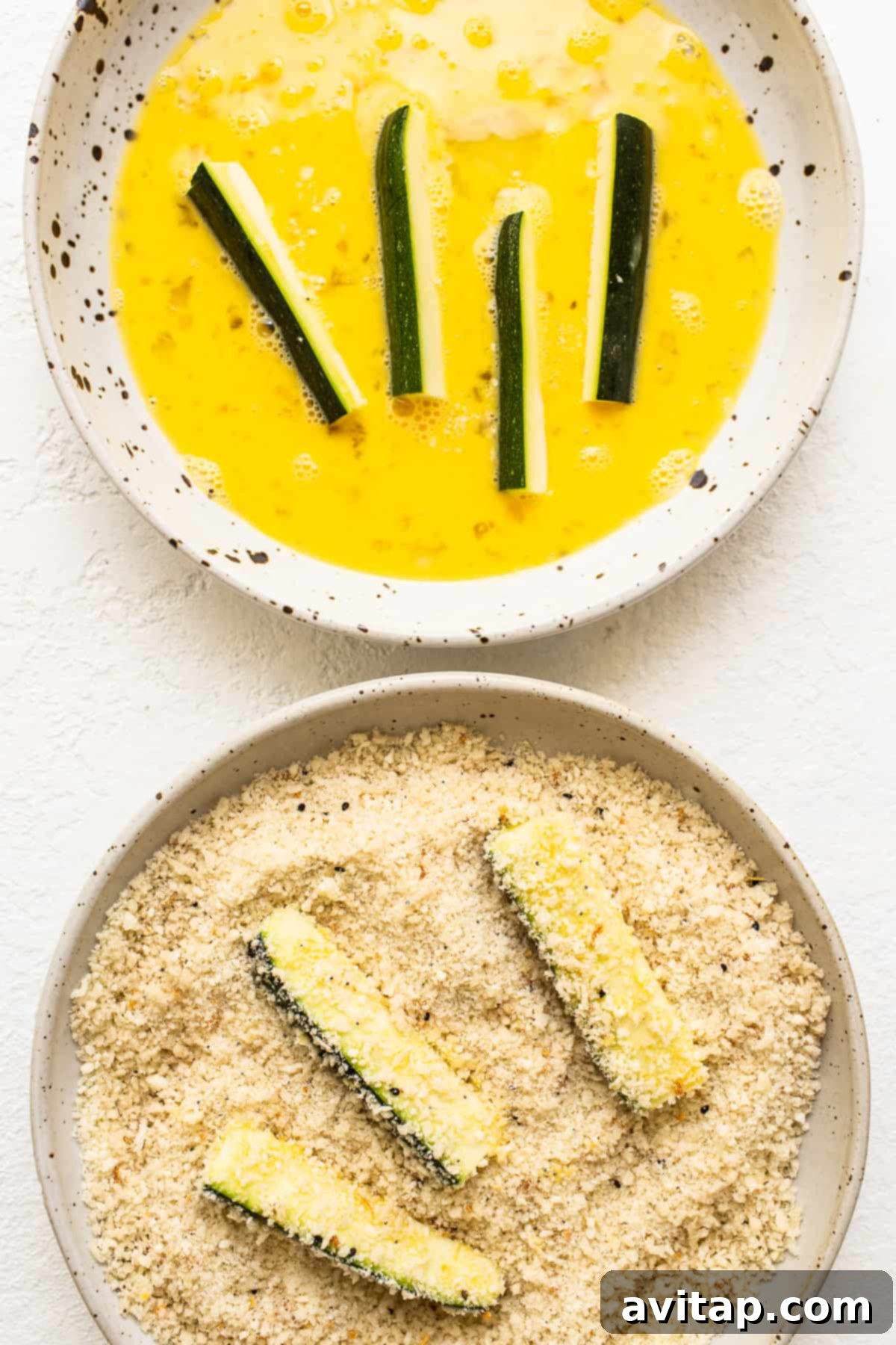 A hand dipping a zucchini spear into a bowl of panko breading mixture.