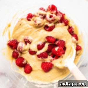 Muffin batter with raspberries folded in, in a mixing bowl, showcasing the vibrant fruit.