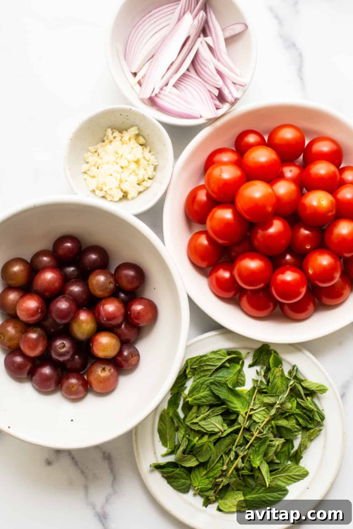 Fresh ingredients laid out on a countertop, including cherry tomatoes, grapes, burrata, garlic, and herbs.