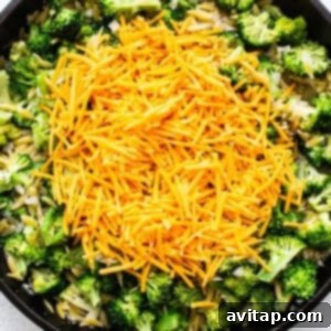 Adding shredded cheddar cheese to the cooked orzo and broccoli in a cast iron skillet.
