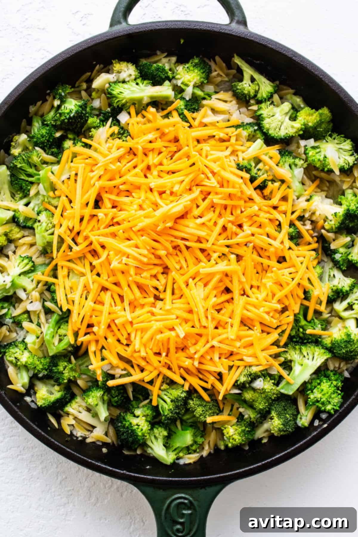 Adding Cheese to Broccoli Orzo Skillet Casserole Shredded cheddar cheese sprinkled generously over cooked broccoli and orzo pasta in a cast iron skillet.