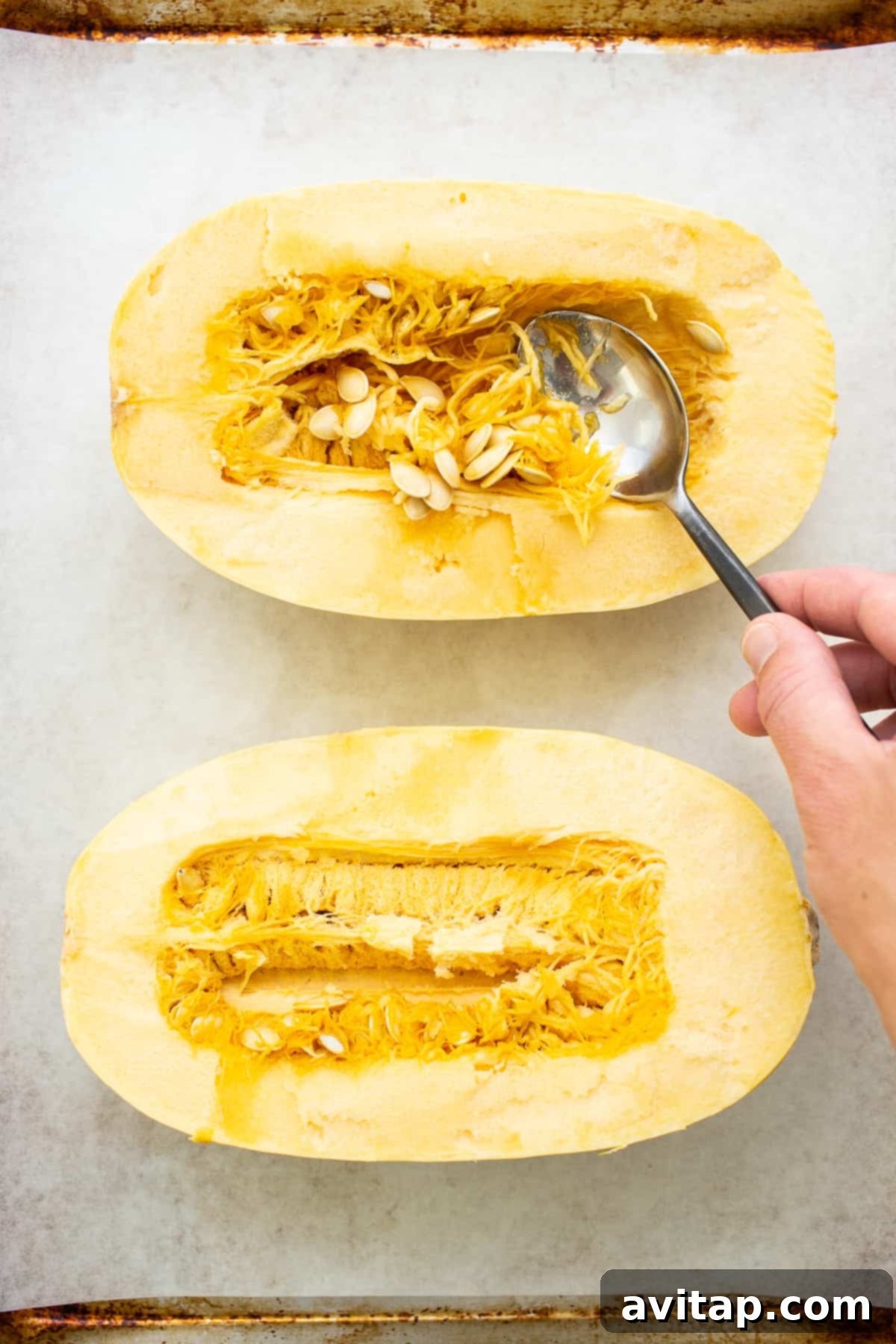 A person expertly spooning out the seeds and stringy innards from a halved spaghetti squash on a kitchen counter.