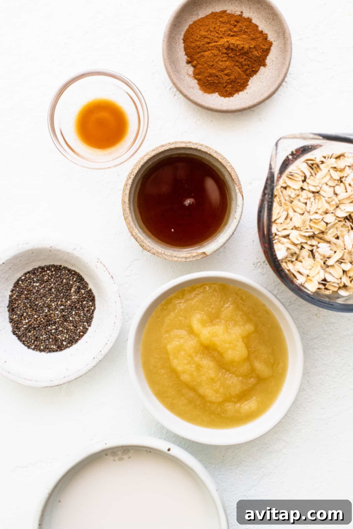 Fresh Ingredients for Apple Cinnamon Oatmeal An inviting assortment of fresh ingredients for apple cinnamon oatmeal, including wholesome rolled oats, unsweetened almond milk, pure maple syrup, smooth applesauce, fragrant vanilla extract, rich ground cinnamon, and tiny chia seeds, all neatly arranged in small bowls.