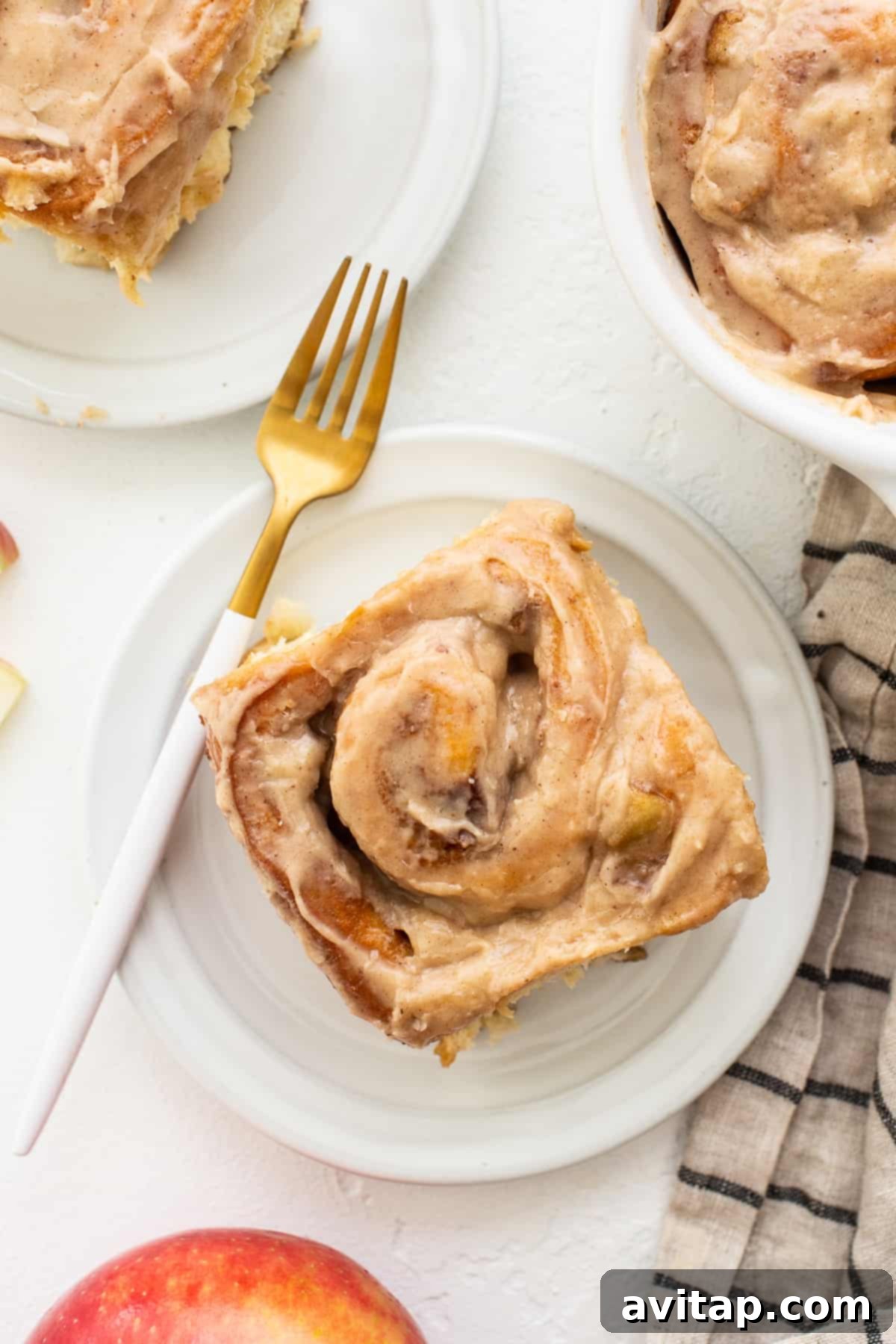 A single apple cinnamon roll, beautifully frosted, resting on a white plate next to a fork, ready to be eaten.
