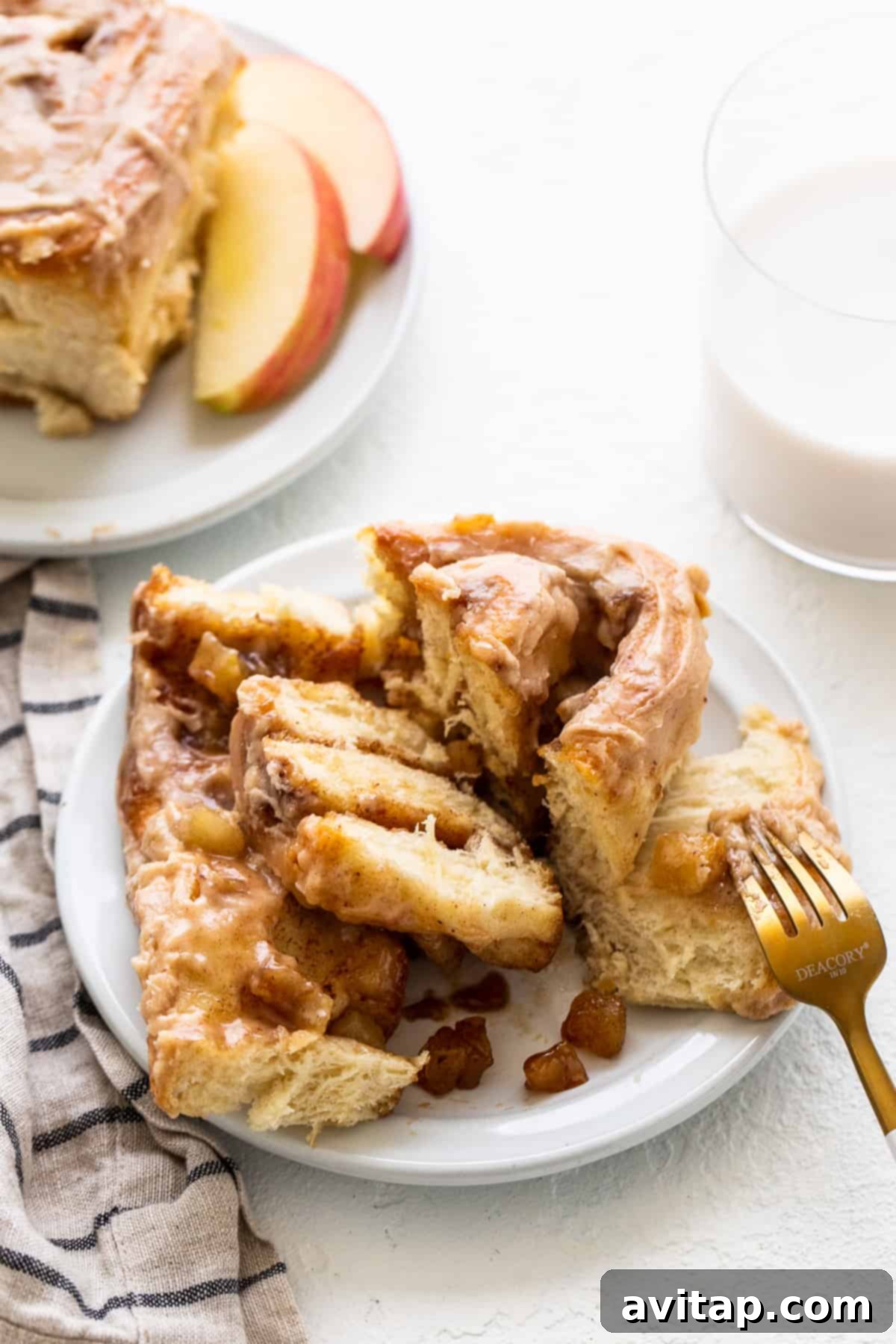 A single apple cinnamon roll, beautifully frosted and garnished, on a white plate with a fork.