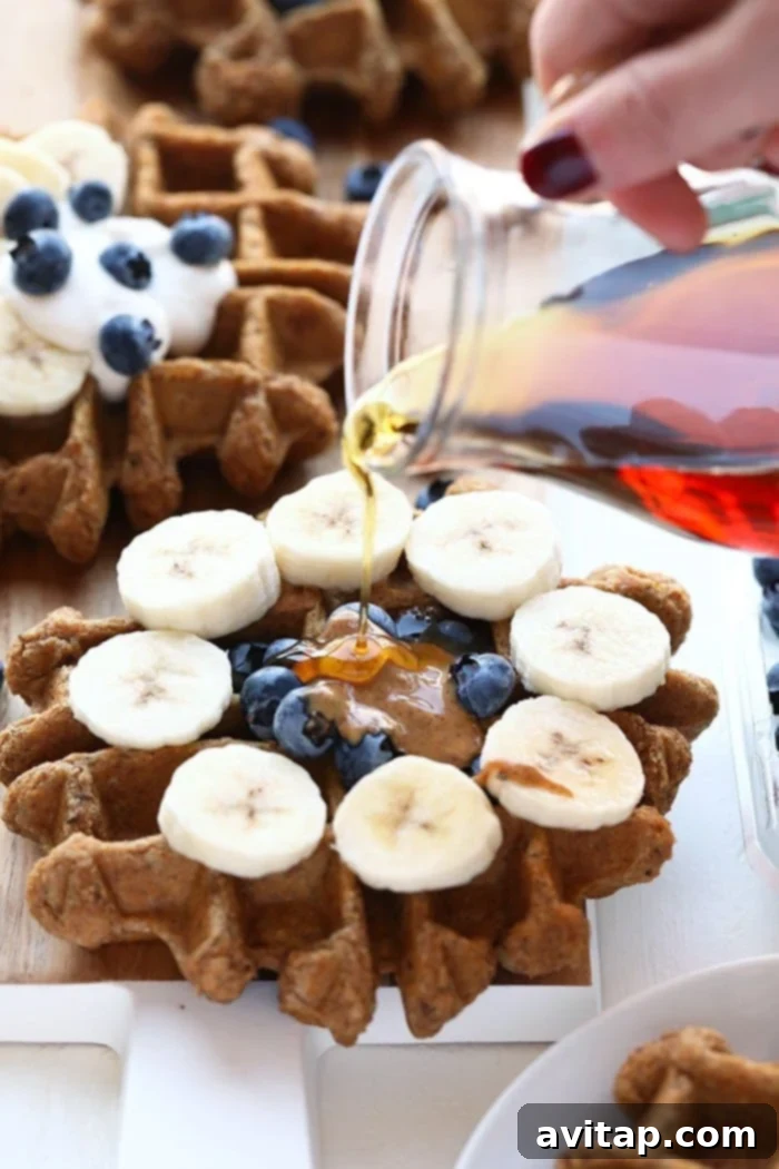A stack of golden-brown vegan almond butter banana bread waffles, topped with fresh banana slices and a drizzle of syrup, on a white plate.