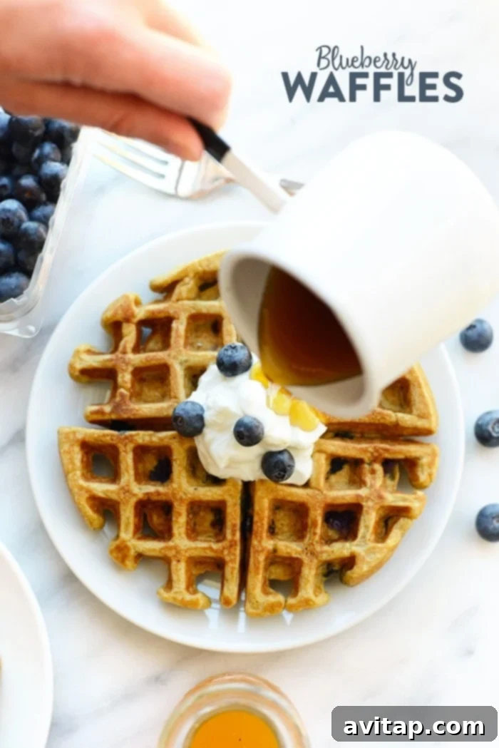 Crispy healthy blueberry waffles, served on a plate and being generously drizzled with maple syrup.