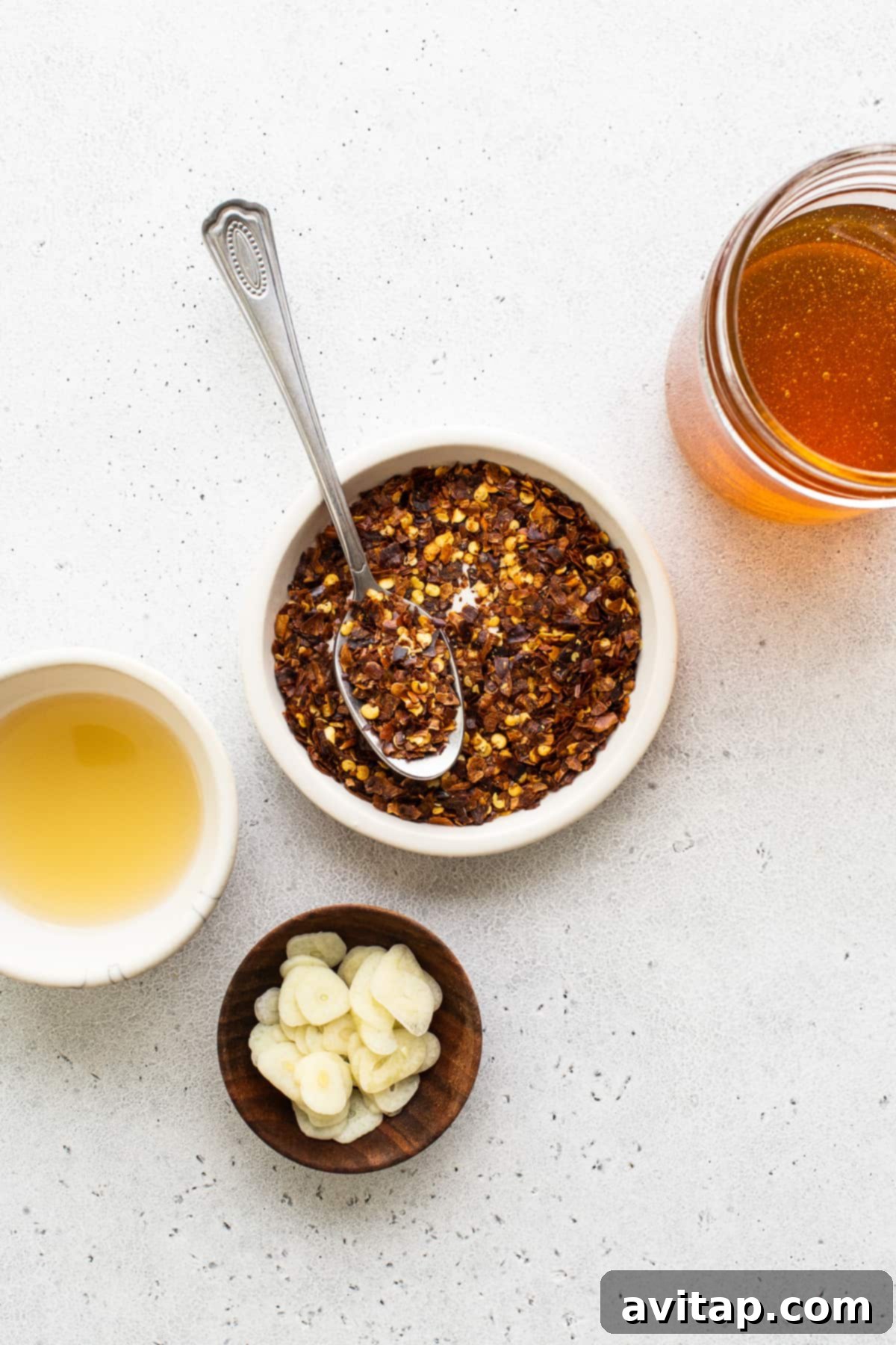 Bowls filled with the four essential ingredients for hot honey sauce: clear filtered honey, bright red pepper flakes, apple cider vinegar, and fresh garlic cloves.