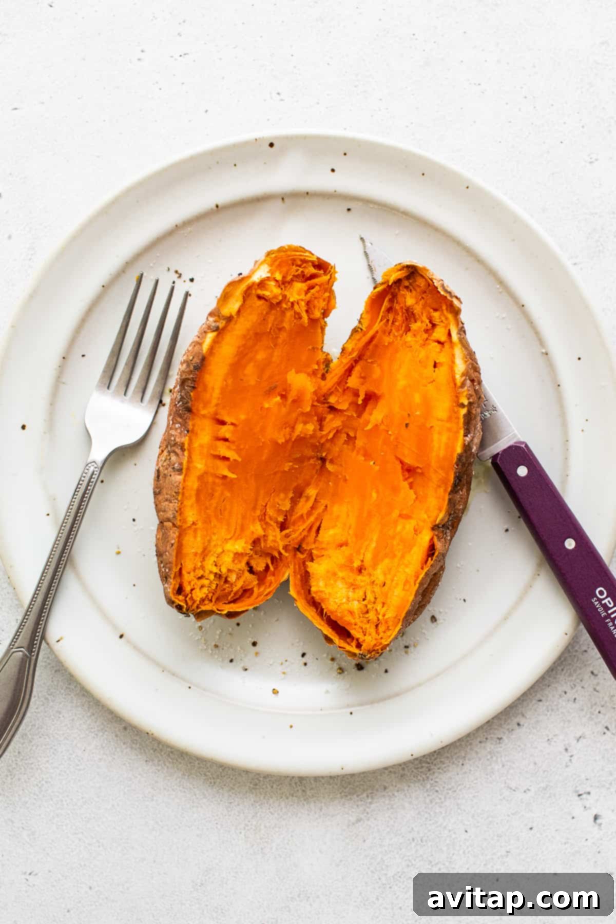 A freshly cooked sweet potato, cut in half on a plate, with a fork gently flaking its tender interior. A pat of butter is melting within.