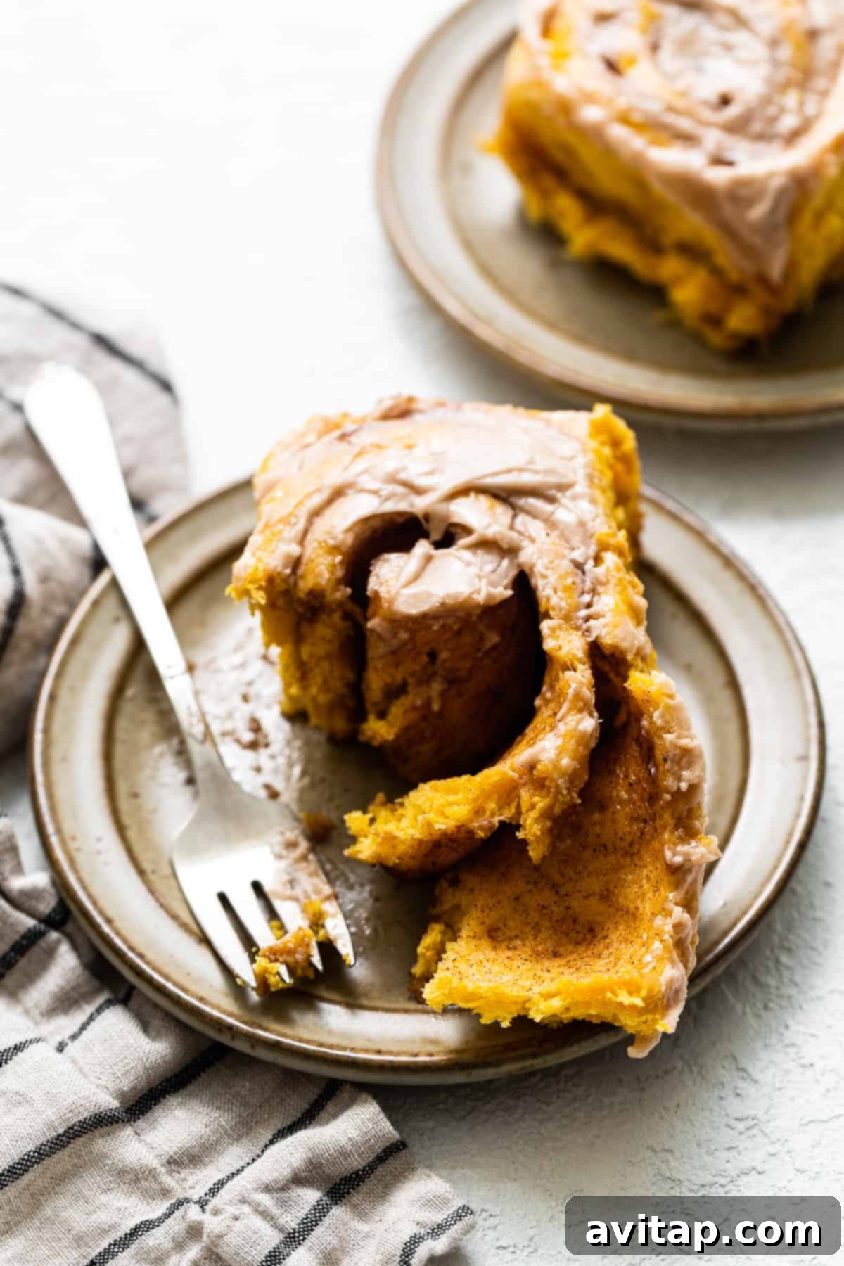 Enjoy a close-up of a delectable pumpkin cinnamon roll, perfectly frosted and ready to be savored. A single frosted pumpkin cinnamon roll on a white plate, showcasing its soft texture and the generous cream cheese topping.